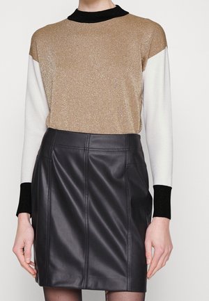 Femme portant un haut à manches longues color block avec des détails dorés, blancs et noirs, associé à une mini-jupe noire en similicuir taille haute.