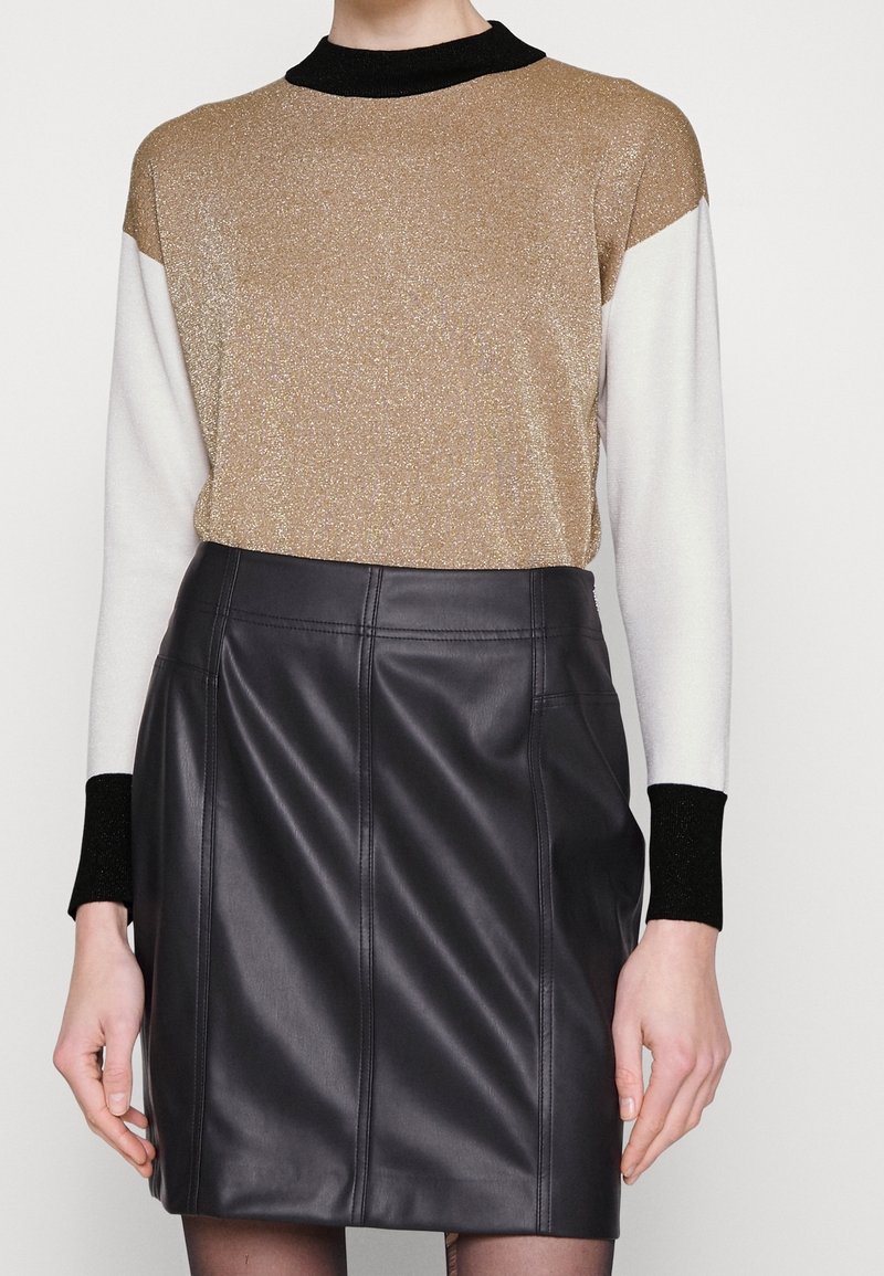 Femme portant un haut à manches longues color block avec des détails dorés, blancs et noirs, associé à une mini-jupe noire en similicuir taille haute.