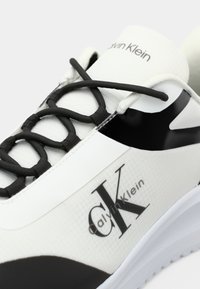 Enfoque próximo de um ténis branco da Calvin Klein com logótipo preto, atacadores pretos e um detalhe preto na lateral.