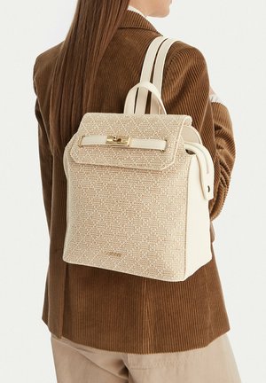 Žena nosící hnědý kordurový blejzr nese texturovaný béžový batoh se zlatou sponou a dvojitými ramenními popruhy.