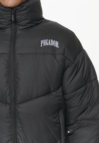 Svart puffjacka med hög krage, dragkedjeöppning och synlig sömnad. Har vit text "PEGADOR" på bröstet och en slät yta.