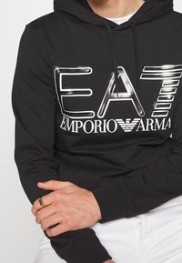 Hombre que lleva una sudadera negra EA7 Emporio Armani con letras blancas, pantalones blancos y un anillo dorado con una piedra verde en la mano izquierda.