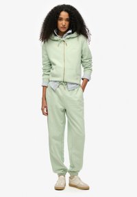 Felpa verde chiaro con cappuccio regolabile, chiusura con zip e polsini a coste, abbinata a pantaloni jogger coordinati. Camicia a righe sotto. Sneakers visibili.