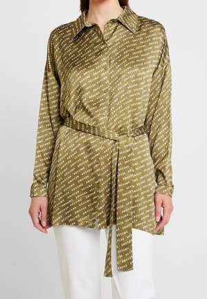 Mujer con blusa túnica verde oliva con botones, patrón diagonal de texto blanco "RÉSUMÉ" y cinturón a juego, combinada con pantalones blancos.