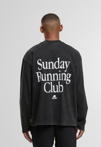 Osoba z krótkimi włosami ubrana w czarną koszulę z długim rękawem, na plecach z napisem "Sunday Running Club", na jednolitym szarym tle.