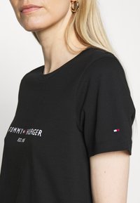 Blondýnka oblečená v černém tričku Tommy Hilfiger s logem a malou vlajkou na rukávu, doplněná zlatými kruhovými náušnicemi.