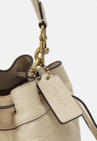 Borsa a secchiello in pelle beige con finitura testurizzata, hardware dorato e un ciondolo rotondo. Presenta un'etichetta con il nome attaccata da una piccola catena.