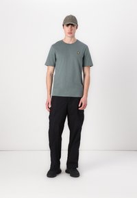 Camiseta de algodón gris con un pequeño logo amarillo, combinada con pantalones cargo negros. El modelo también lleva una gorra gris y zapatillas negras.
