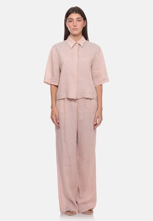 Donna in piedi che indossa una camicia rosa chiaro larga con maniche corte e pantaloni a gamba larga abbinati su sfondo bianco.