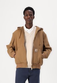 Veste à capuche marron avec fermeture éclair à l'avant, grandes poches, poignets côtelés et un patch logo Carhartt. Portée par-dessus un pull de couleur claire.