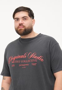 Tumši pelēks kokvilnas t-krekls ar īsām piedurknēm, ar sarkanu grafisku tekstu: "Originals Studio Artist Collective JACK&JONES Gallery Tour."