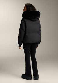 Žena v čiernej nadýchnutej bunde s veľkou kapucňou lemovanou kožušinou, čiernymi priliehavými nohavicami a čiernymi platformovými topánkami, stojaca pred neutrálnym pozadím.