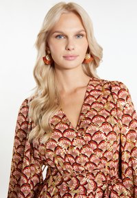 Rochie înflorată cu un model roșu și auriu, cu mâneci lungi, decolteu adânc în V și talie cu șnur. Cercei rotunzi inspirați din ceramică, cu franjuri.