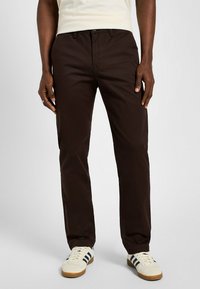 Brązowe bawełniane spodnie o dopasowanym kroju, z zamkiem błyskawicznym, szlufkami na pasek i pojedynczymi przednimi kieszeniami. Gładka faktura i minimalna liczba szwów.