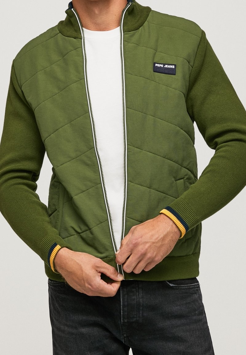 Grön vadderad zip-jacka med hög krage, kontrasterande ribbade manschetter i marinblått och gult, bärs över en vit skjorta. Svarta jeans syns.