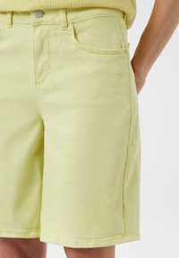 Jasnożółte szorty dżinsowe o prostym kroju, pięć kieszeni, subtelne przeszycia i metalowy guzik w pasie. Gładka tekstura.