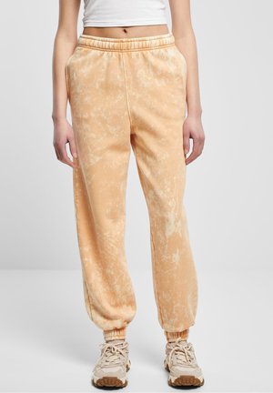 Mujer con pantalones jogger beige teñidos tipo tie-dye con cintura y puños elásticos, top corto blanco y zapatillas beige, de pie sobre fondo gris.