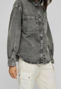 Chemise en jean grise avec deux poches poitrine, fermeture à boutons et manches retroussées, associée à un pantalon cargo crème avec poches plaquées.