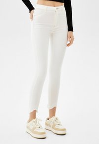Pantalones vaqueros blancos ajustados de mezclilla con dobladillo deshilachado, que cuentan con una cintura alta, cierre de botón y costuras en contraste amarillas. Combinados con zapatillas de deporte beige.
