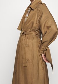 Długi brązowy trencz z lekkiego materiału, z paskiem w talii, dużymi guzikami i podwiniętymi rękawami, które odsłaniają wzorzysty wnętrze.