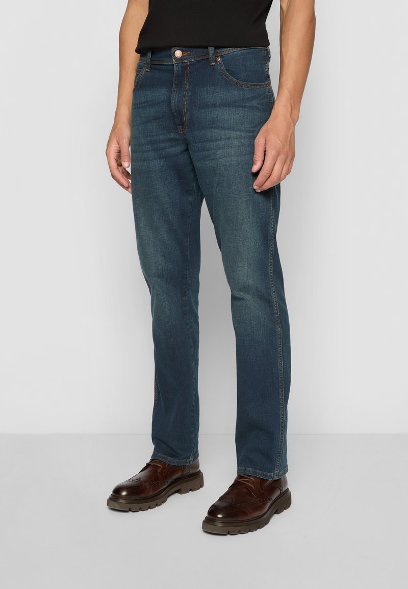 Man wearing faded dark blue jeans and brown leather brogue shoes, standing against a plain light gray background.