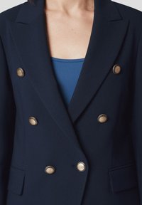 Granatowa, dwurzędowa marynarka z złotymi guzikami, klapami i dwoma przednimi kieszeniami. Gładka tkanina i dopasowany krój.