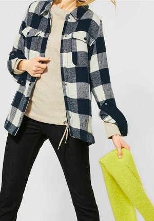 Osoba ubrana w beżowy sweter, czarno-białą kratowaną kurtkę, czarne spodnie, trzymająca jasny żółty szalik w jednej ręce.