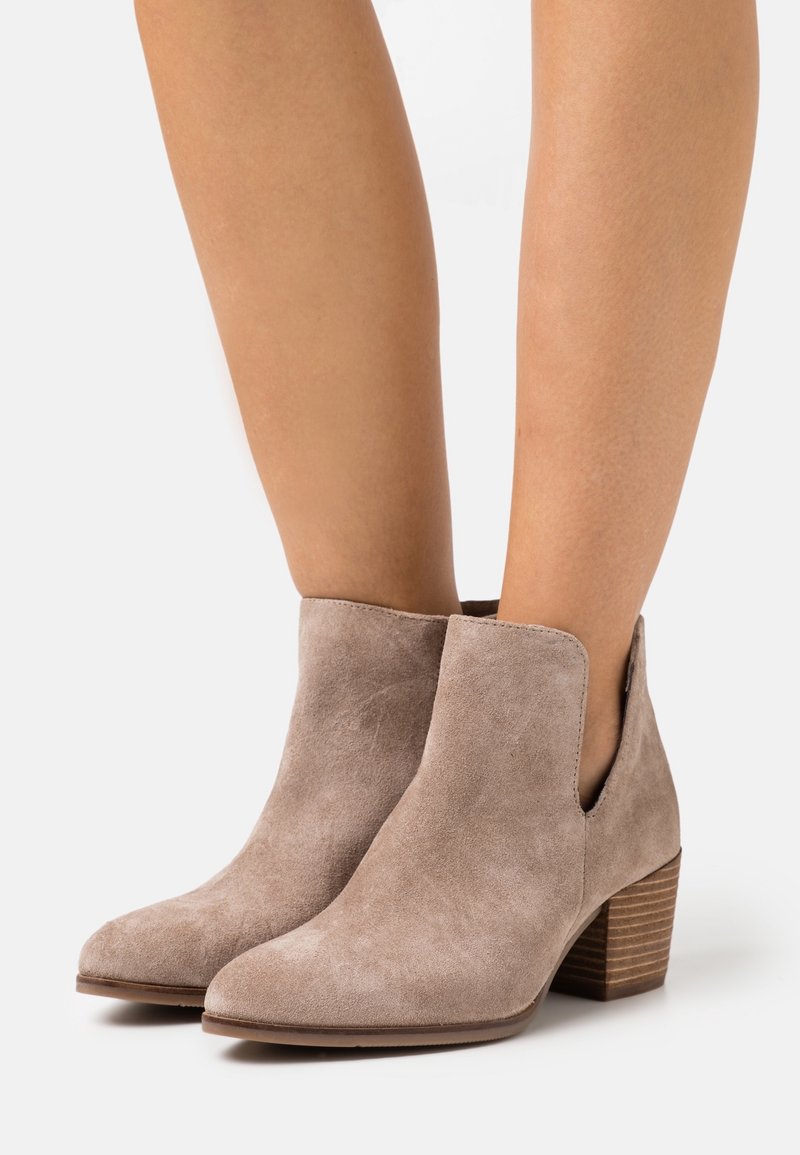 Bottines en daim taupe avec talons blocs empilés portées sur des jambes bronzées sur fond blanc.
