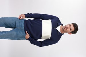 Homme portant un pull marine et blanc à demi-fermeture éclair et un jean bleu, debout les mains dans les poches devant un fond blanc uni.