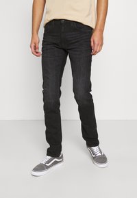 Homme portant un jean slim noir et des baskets grises avec des lacets blancs, se tenant devant un fond gris clair uni.