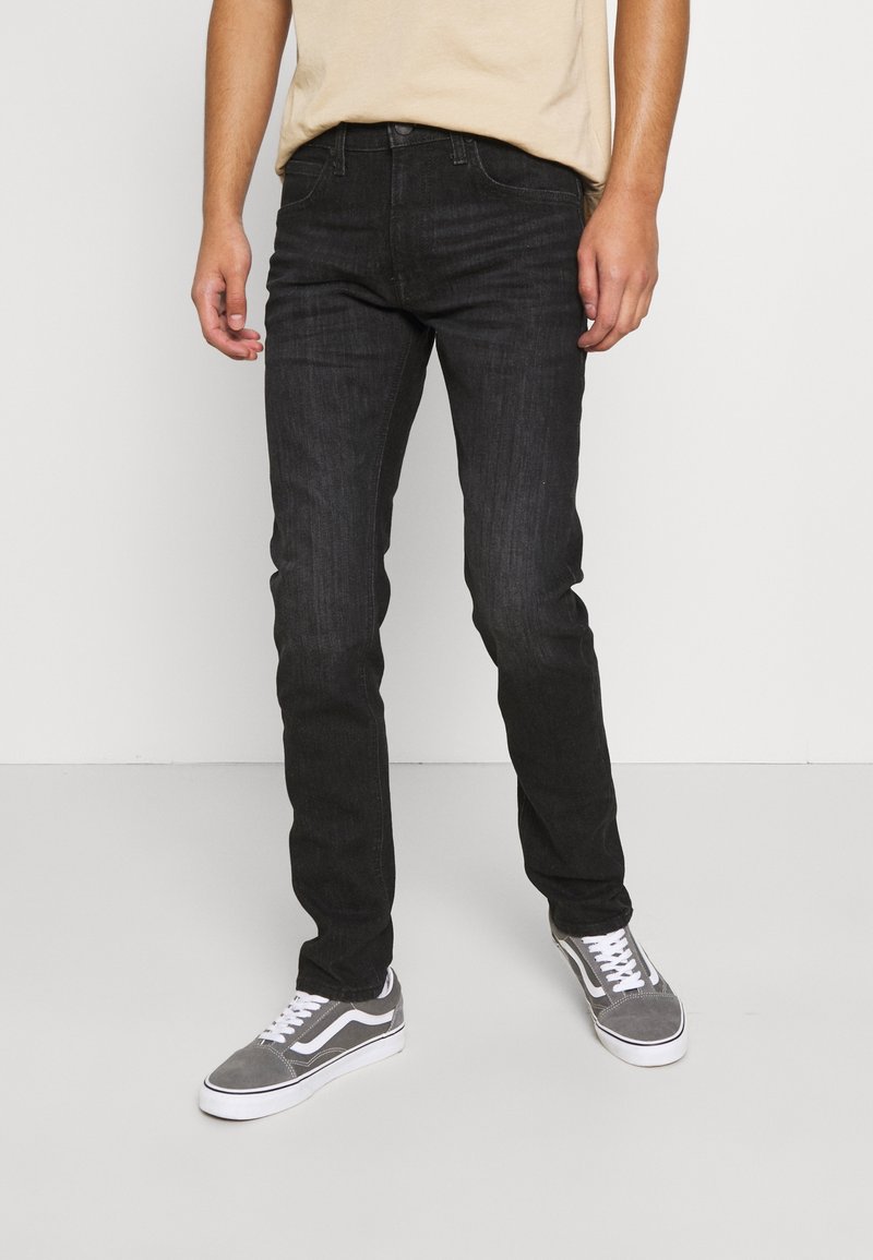 Homme portant un jean slim noir et des baskets grises avec des lacets blancs, se tenant devant un fond gris clair uni.