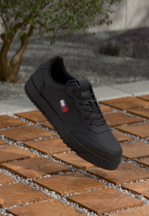 Schwarzer Sneaker mit Schnürsenkeln und Tommy Hilfiger-Logo, der über quadratischen Holzdielen mit Kieselsteinen dazwischen schwebt, im Freien in der Nähe eines Baumes und von Steinstufen.