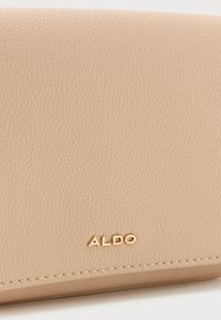 Beige teksturēta ādas maks ar zelta ALDO logotipu un redzamām šuvēm apakšējā malā uz balta fona.