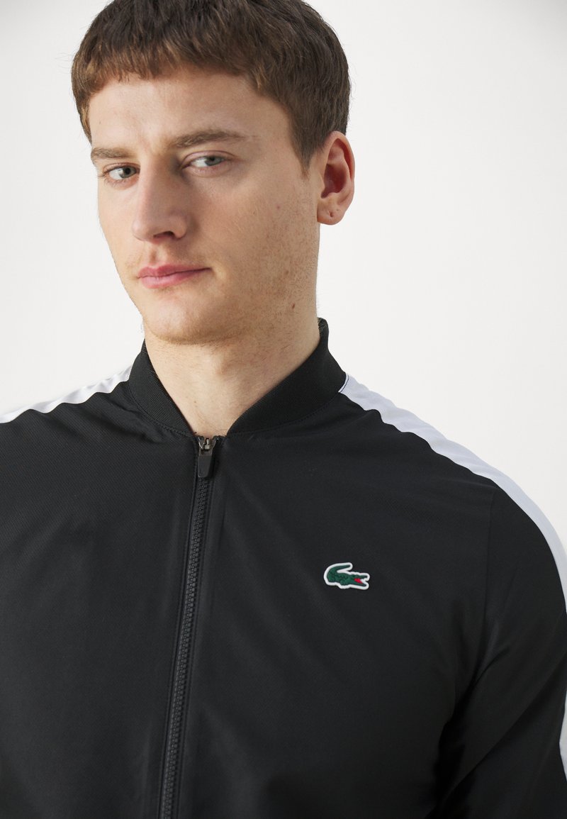 Giacca nera con zip e colletto alto, con strisce bianche sulle spalle e un logo di coccodrillo verde sul petto. Texture liscia.