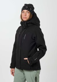 Vrouw met een zwarte capuchonjas en een zwart gemêleerde muts, staand met één hand in haar zak, kijkt iets naast zich.