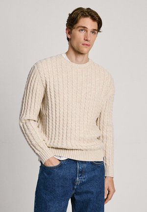 Suéter de punto acanalado beige con cuello redondo y mangas largas, combinado con jeans de mezclilla azul oscuro. La textura es suave con patrones de trenzado visibles.