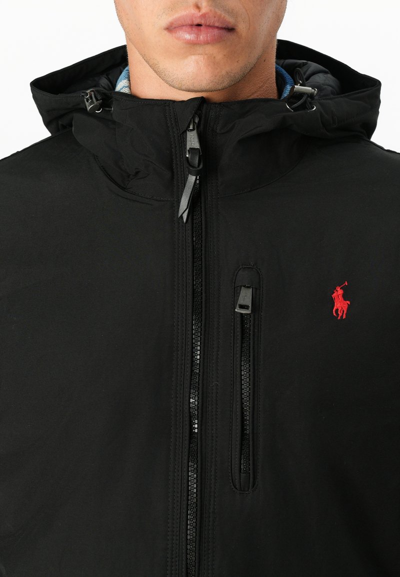 Czarna kurtka z kapturem z przednim zamkiem, zamykanymi kieszeniami i czerwonym logo polo. Materiał wydaje się gładki o matowym wykończeniu.