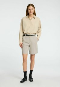 Beige langærmet skjorte, grå skræddersyede shorts, sort bælte med guldfarvet spænde, sorte loafers og sorte sokker på en ensfarvet baggrund.
