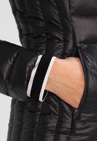 Crni puffa kaput s kvadratnom teksturom, elegantnim dizajnom, zatvorenim džepom i kontrastnim bijelim i crnim prugastim manžetama koje vire.