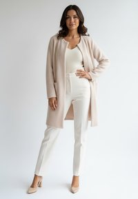 Jasnoróżowy długi płaszcz o miękkiej fakturze, z otwartym przodem bez guzików, noszony na białym prążkowanym topie i białych, dopasowanych spodniach, uzupełniony beżowymi szpilkami.