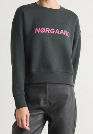 Personne portant un sweat-shirt vert foncé avec un texte rose "NØRGAARD" et un pantalon en cuir noir, debout devant un fond clair uni.