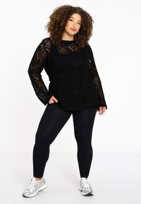 Zwarte, gebreide trui met lange mouwen met opengewerkte patronen. Combinatie met zwarte leggings en zilveren sneakers.