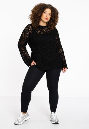 Zwarte, gebreide trui met lange mouwen met opengewerkte patronen. Combinatie met zwarte leggings en zilveren sneakers.