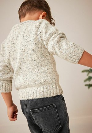 Enfant portant un pull en maille beige tacheté et un jean gris foncé, vu de dos avec les bras légèrement étendus devant un fond neutre.