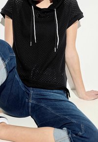 Czarna bluzka z krótkim rękawem o siatkowej strukturze, z kapturem i białymi sznurkami. Połączona z niebieskimi dżinsami, co tworzy casualowy styl.