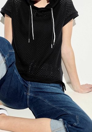 Haut noir à manches courtes avec un design en maille, une capuche et des cordons blancs. Associé à un jean en denim bleu, montrant un style décontracté.