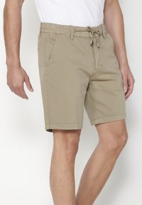 Vīrietis ģērbjas bēšos līdz ceļgaliem šortos ar pievilkšanas jostu un sānu kabatām, kas kombinēti ar balu T-kreklu.