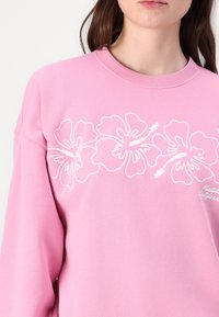 Roza dukserica s bijelom vezenom hibiskus cvijetom preko prsa, s okruglim izrezom i dugim rukavima. Mekana, teksturirana tkanina.