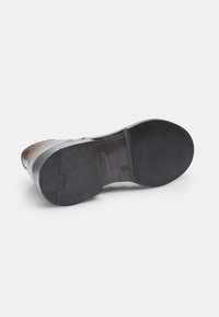 Suela de goma negra de un zapato, con un diseño plano, superficie texturizada y una impresión de logo. El material superior presenta una sección de cuero marrón.