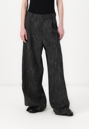 Pantalons larges en tissu noir avec un motif texturé et subtil. La taille est plate et présente des poches de chaque côté. Chaussures noires visibles.